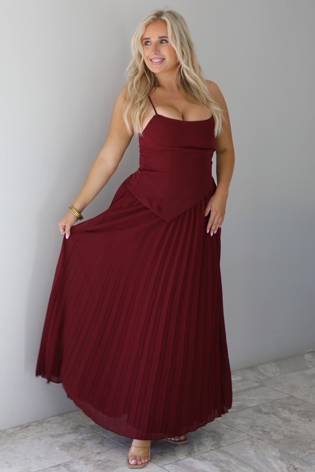 A woman wearing a wine colored formal maxi dress with a fitted, drop waist design with a crimped floral maxi skirt, standing indoors against a white wall with white marble flooring. 