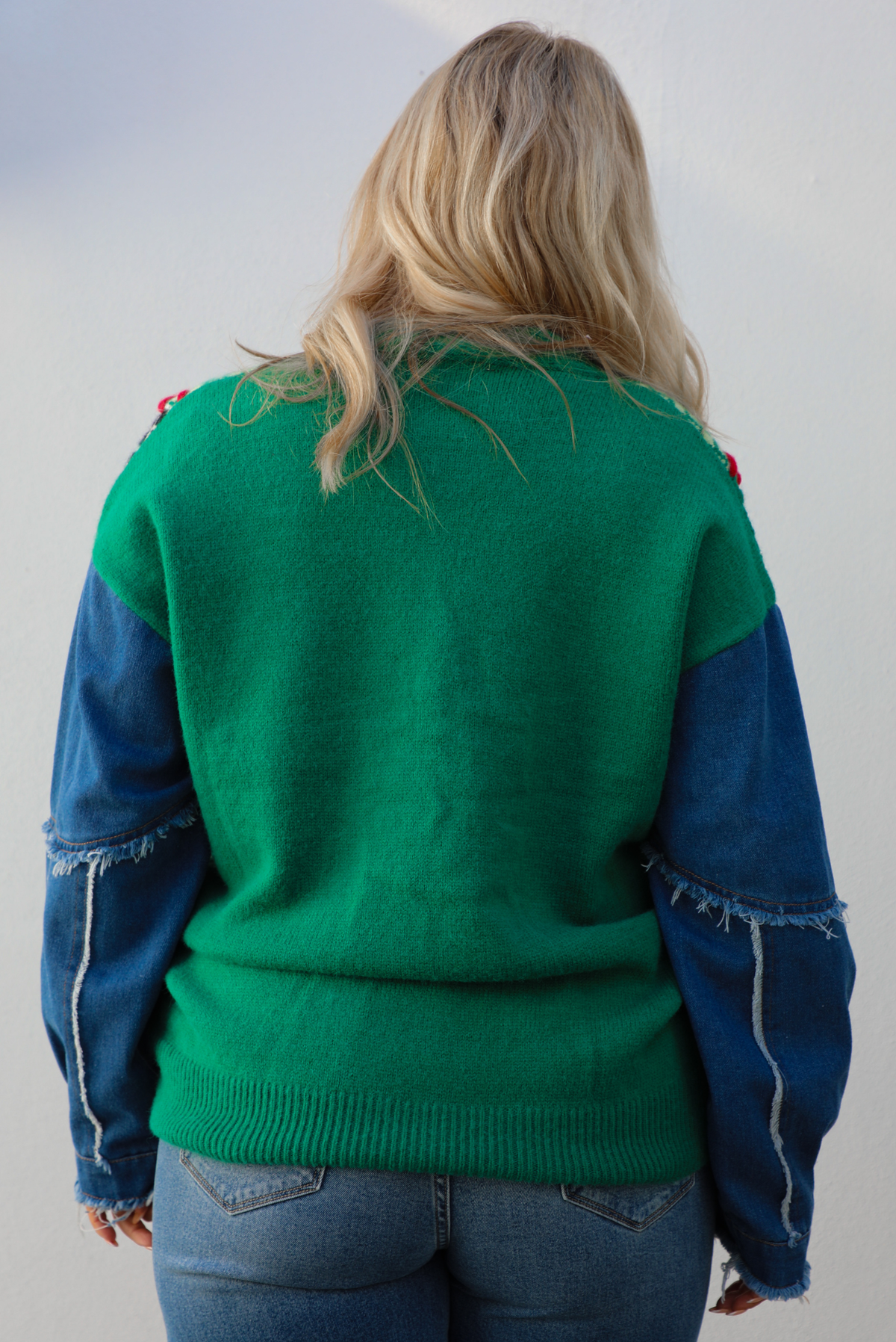 Woman model wearing a green and multi-colored cardigan with denim sleeves, and medium wash denim jeans. The photo shows up a close up of the back of the cardigan and its full length, and the frayed, raw hem details on the sleeves. The photo is taken against a white wall outdoors.