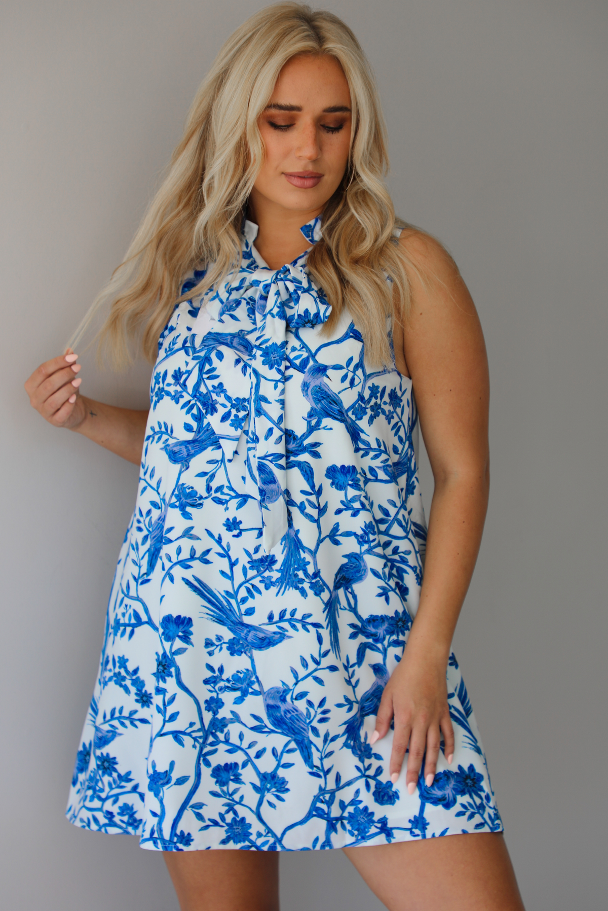 Woman wearing a white and blue print dress with a tree branch and bird pattern throughout indoors against a white wall.
