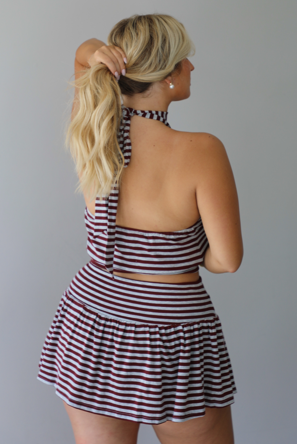 A woman shown from the back wearing a burgundy and grey striped two piece set featuring a high-neck cropped top and mini skirt, high scrunched white socks, and brown leather loafers, standing indoors against a white wall.