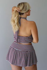 A woman shown from the back wearing a burgundy and grey striped two piece set featuring a high-neck cropped top and mini skirt, high scrunched white socks, and brown leather loafers, standing indoors against a white wall.