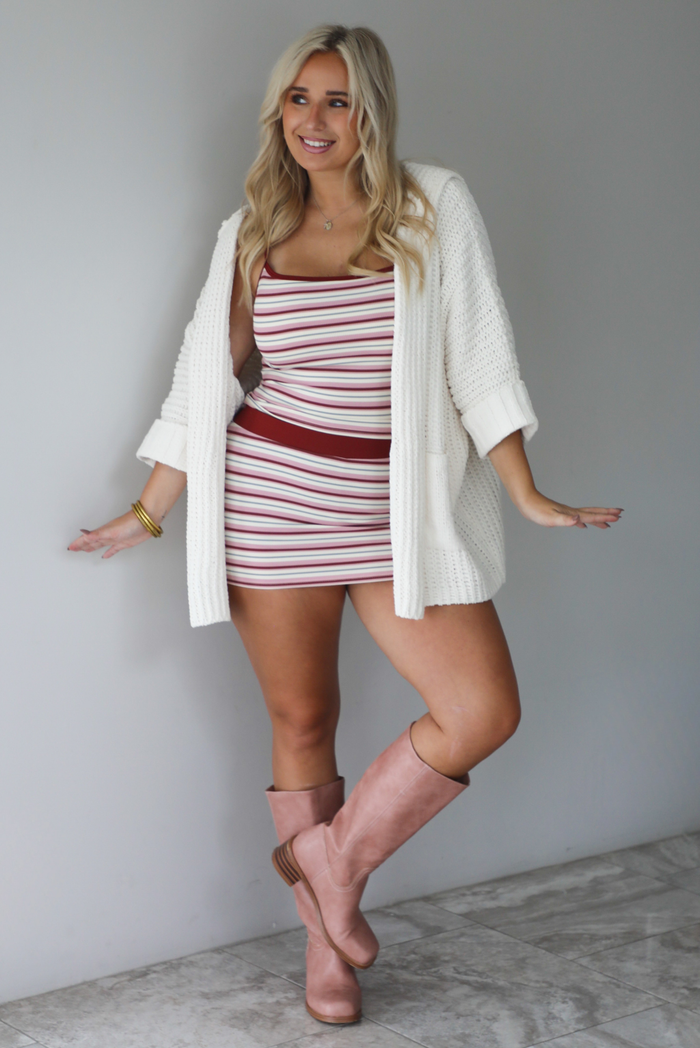A woman wearing a cream colored oversized cardigan worn over a striped two piece set, paired with knee high pink boots, standing indoors against a white wall with white marble flooring. 