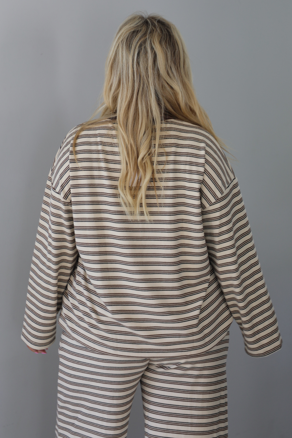 A woman shown from behind wearing a sand colored matching two piece set with a brown striped pattern throughout featuring an oversized long sleeved top and oversized slouchy wide leg pants, standing indoors against a white wall. The photo shows a closeup of the top from behind, untouched to show the full length. 