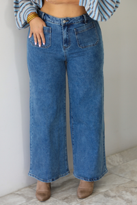 A woman wearing blue wide-leg denim jeans with front patch pockets, paired with a striped long-sleeve top and tan pointed-toe booties, standing indoors against a white wall with white marble flooring. 