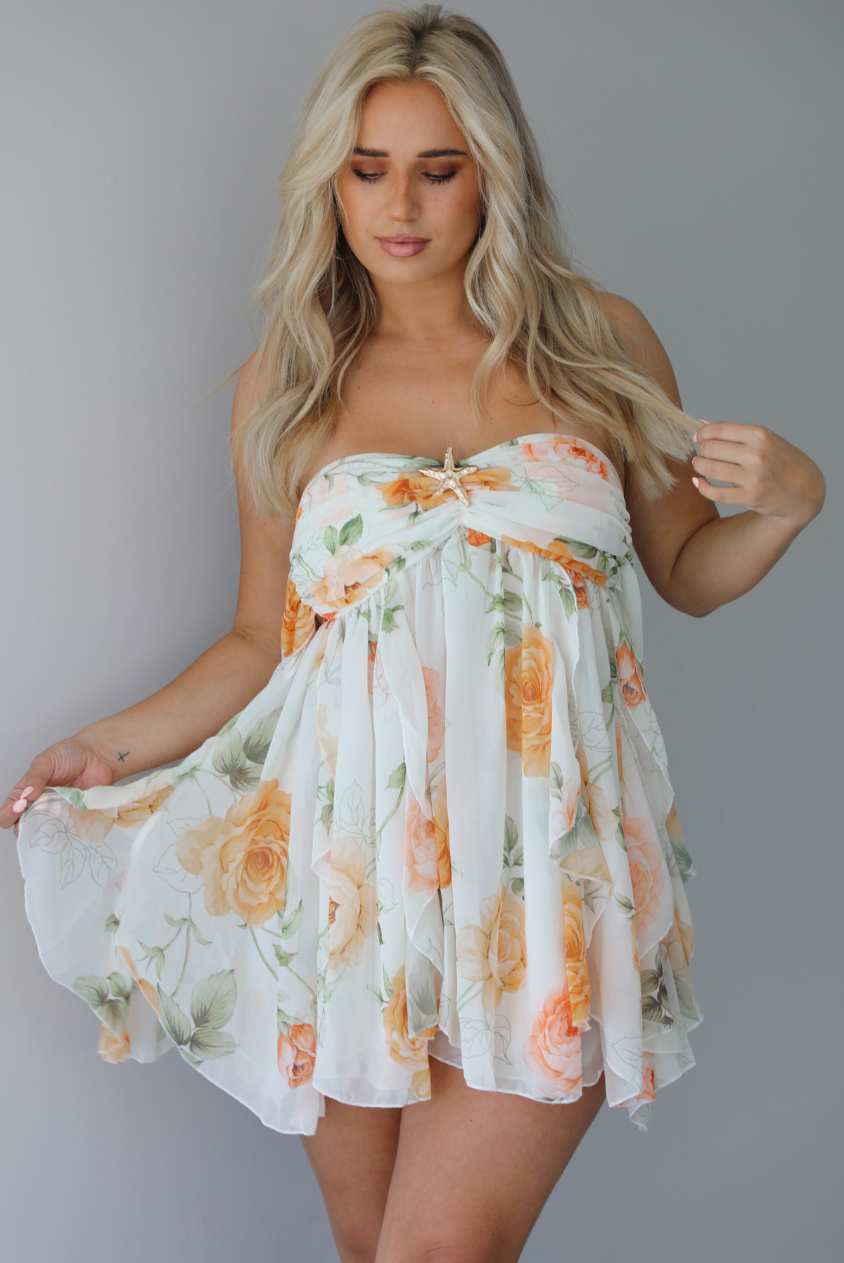 Woman wearing a white, strapless, babydoll cut dress that has an orange and green floral print pattern and flowy skirt indoors against a white wall.