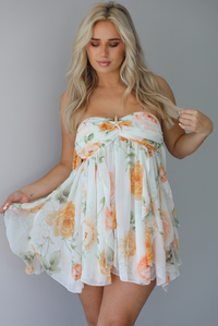 Woman wearing a white, strapless, babydoll cut dress that has an orange and green floral print pattern and flowy skirt indoors against a white wall.