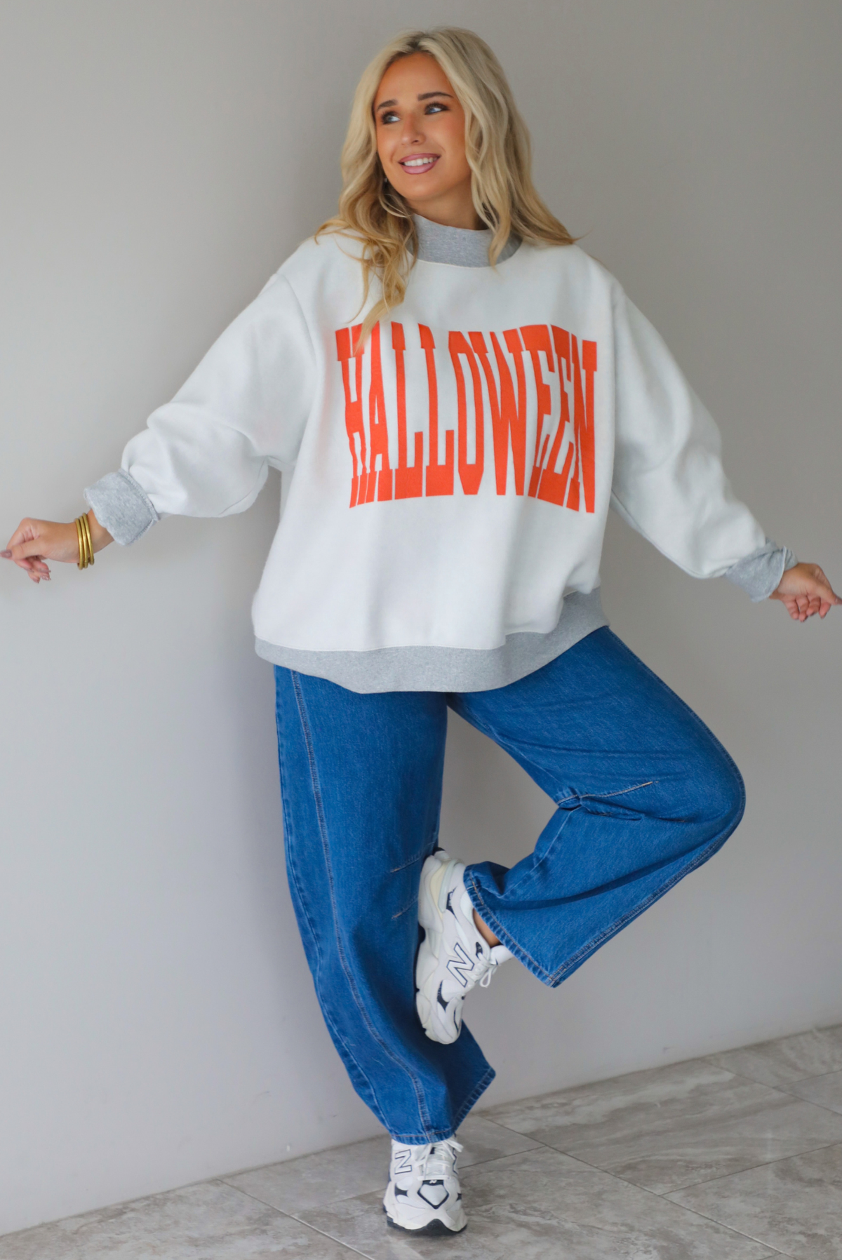 A woman wearing a light grey and dark grey trim oversized sweatshirt that says "HALLOWEEN" in collegiate font on the front, with jack-o-lantern details on the rolled up sleeves, paired with dark wash denim jeans, standing indoors against a white wall with white marble flooring. 