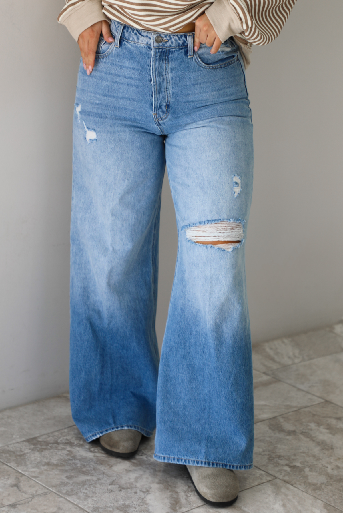 Woman wearing high waisted medium wash denim that have multiple button closures, functional belt loops, functional front and back pockets, and distressed details throughout with a flared hem. The photo is taken in front of a white wall and white marble flooring.