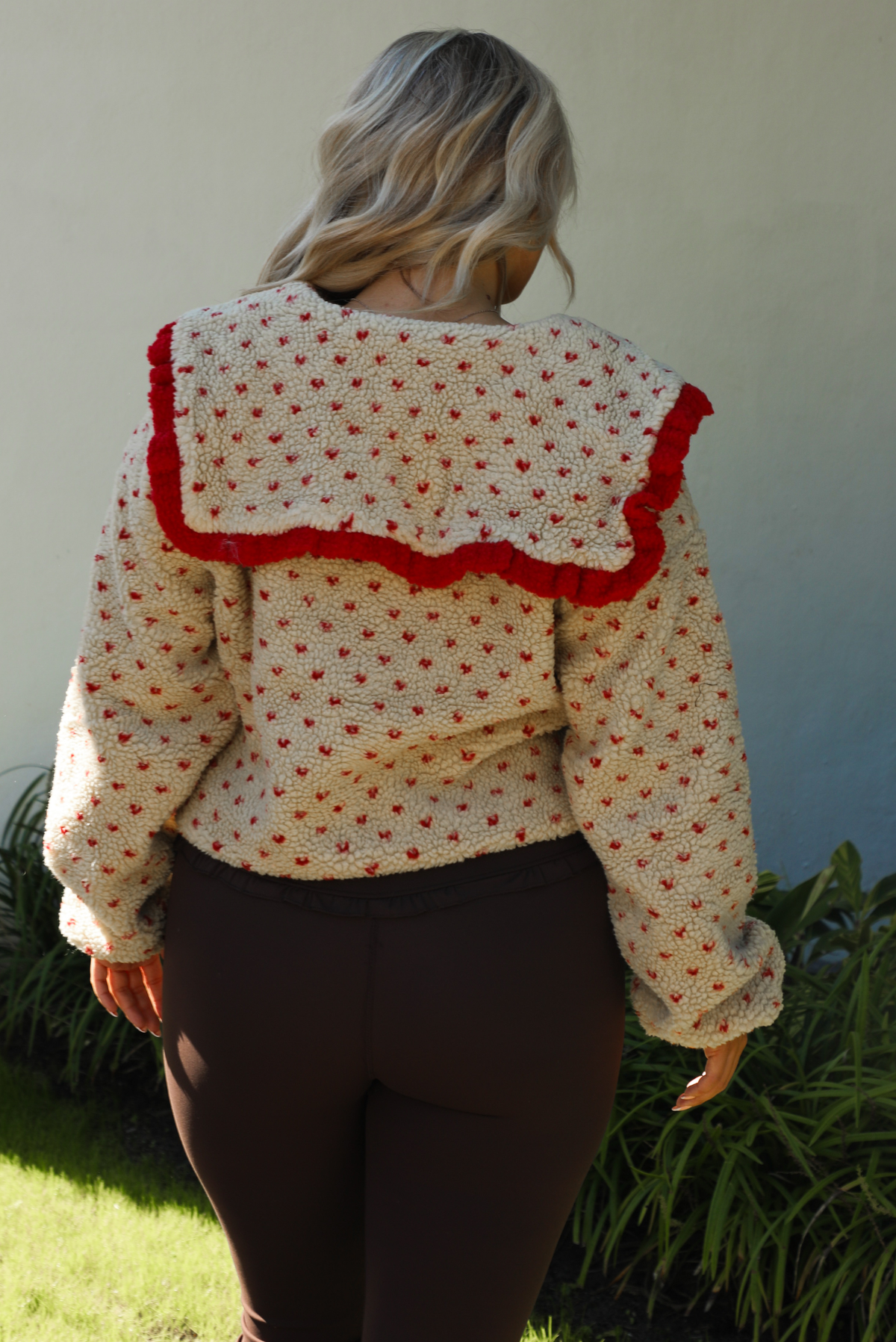 Woman model wearing a tan and red sherpa cardigan with a collared neckline and red trim with dark brown ahtleisure pants. The photo is taken outdoors with a white wall and greenery in the back. Photo showcases the back with the collar detail. 