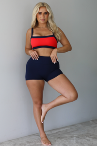 Woman wearing. navy and red sports bra with navy high waisted shorts indoors against a white wall with white marble flooring. 