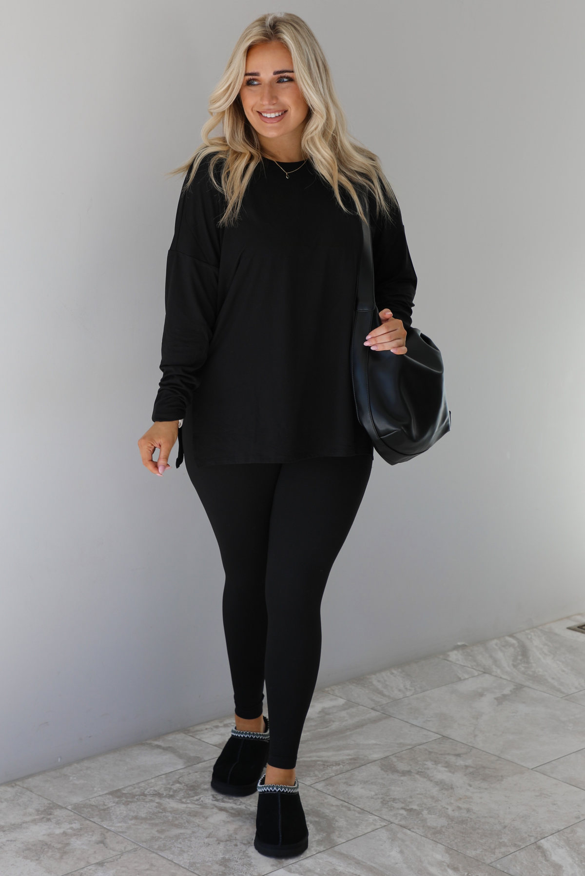 Woman wearing a black two piece set that features an oversized long sleeved top and fitted leggings. The set is paired with a black vegan leather shoulder bag and black slide on slippers. The photo is taken indoors against a white wall with white marble flooring.