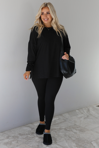 Woman wearing a black two piece set that features an oversized long sleeved top and fitted leggings. The set is paired with a black vegan leather shoulder bag and black slide on slippers. The photo is taken indoors against a white wall with white marble flooring.