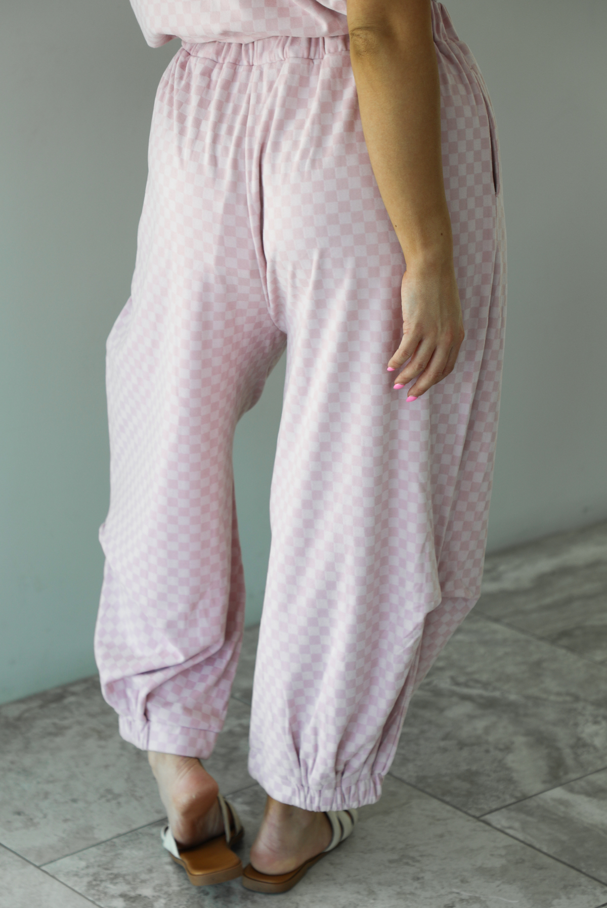 Woman wearing a pink checkered set featuring a slouchy top and pants. The photo is taken indoors against a white wall and shows a closeup of the back of the pants, which are high waisted, have an elastic waistline, functional pockets on either side, have an oversized fit, and cinch slightly with elastic at the hem of the pants.