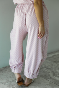 Woman wearing a pink checkered set featuring a slouchy top and pants. The photo is taken indoors against a white wall and shows a closeup of the back of the pants, which are high waisted, have an elastic waistline, functional pockets on either side, have an oversized fit, and cinch slightly with elastic at the hem of the pants.
