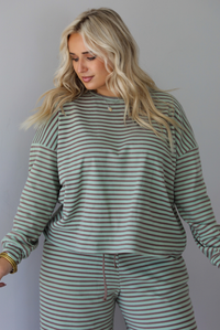 A woman wearing a pale green and brown striped two piece set featuring an oversized long sleeved top and high waisted, wide leg pants, standing indoors against a white wall.