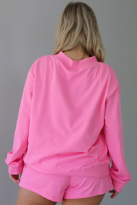 Woman wearing a hot pink two piece set featuring an oversized sweatshirt and high waisted shorts. The photo is taken indoors against a white wall and shows a closeup of the back of the top of the set, which is untucked to show the full length. 