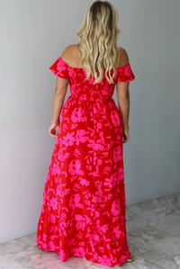 A woman shown from behind modeling a bright red off-the-shoulder maxi dress with a vibrant pink floral pattern, standing indoors agains a white wall with white marble flooring. 