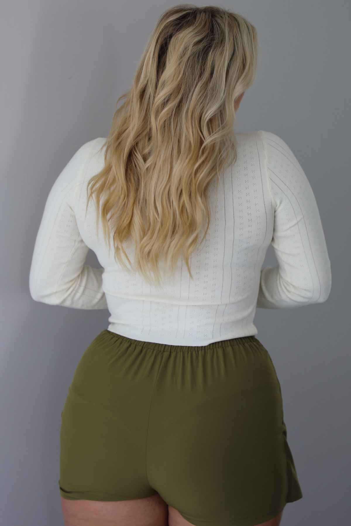A woman shown from behind wearing a cream colored long sleeved top with an olive green skort, indoors against a white wall. 
