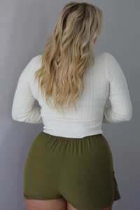 A woman shown from behind wearing a cream colored long sleeved top with an olive green skort, indoors against a white wall. 