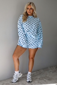 A woman wearing a blue two piece set with a navy polka dot pattern throughout, featuring a sweatshirt and high waisted sweat-shorts, paired with chunky sneakers and high ankle socks, shown indoors against a white wall with white marble flooring. 