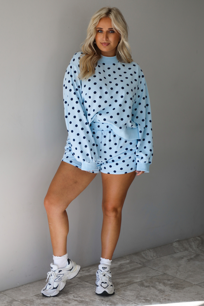 A woman wearing a blue two piece set with a navy polka dot pattern throughout, featuring a sweatshirt and high waisted sweat-shorts, paired with chunky sneakers and high ankle socks, shown indoors against a white wall with white marble flooring. 