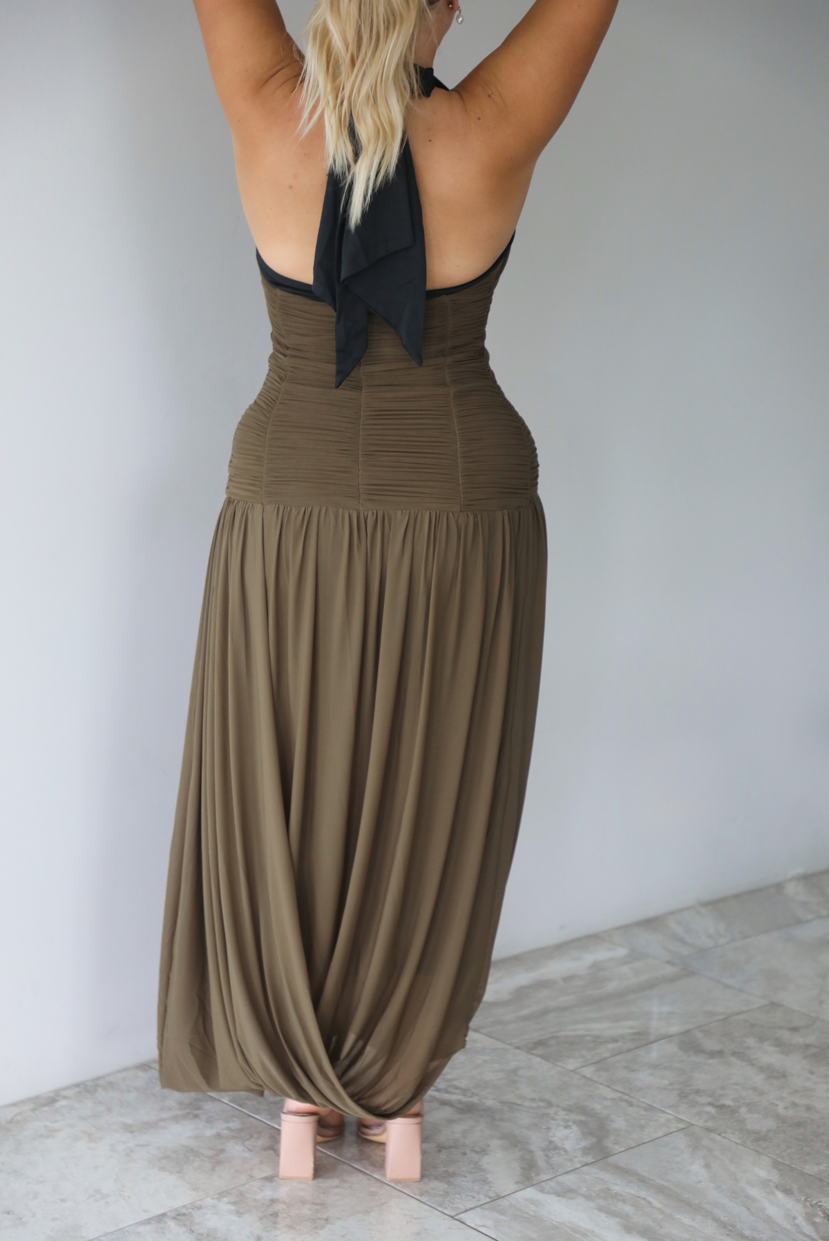 A woman shown from behind wearing an olive maxi dress with black trim detailing at the v-halter neckline, has a fitted bodice and drop waist design, with a maxi-length flowy skirt, standing indoors against a white wall with white marble flooring. The back of the dress has a halter tie neckline. 