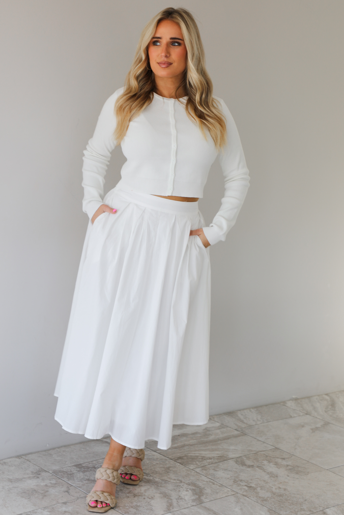 Woman wearing a white two piece set featuring a long sleeved cardigan and a high waisted midi length skirt. The photo is taken indoors against a white wall with white marble flooring. 