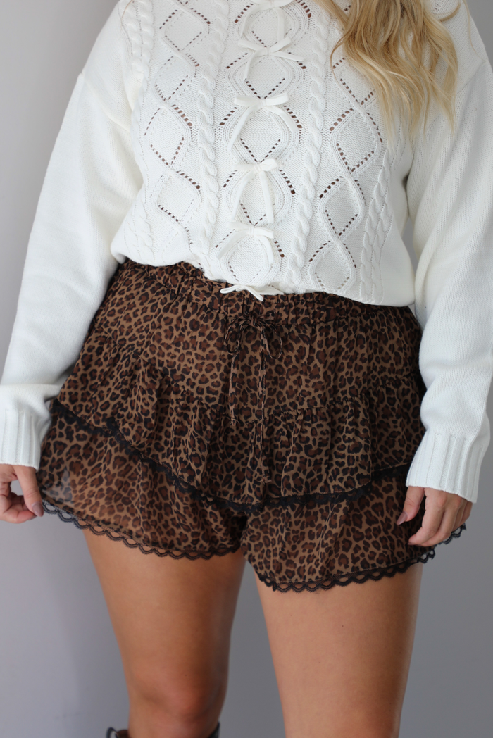 A closeup of a woman shown from the waist down wearing a white sweater with leopard shorts with black lace trim detailing, indoors against a white wall.