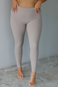 A woman modeling a matching tan activewear set consisting of a sports bra and high-waisted leggings with white stitching details, standing indoors against a white wall.