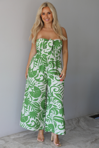 Woman wearing a strapless jumpsuit with green leaf pattern indoors against a white wall with white marble flooring. 