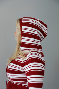 A woman shown from the side modeling a matching red and white striped knit set with the hoodie on the top up, has a front zipper, drawstring hood, standing indoors against a white wall.