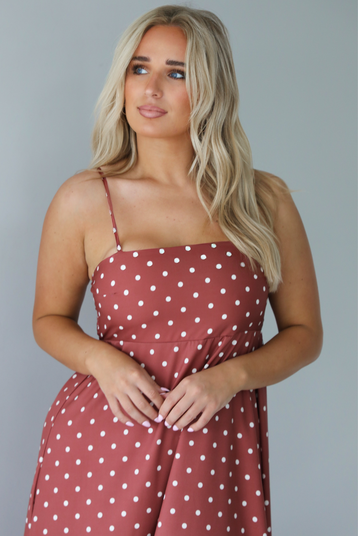 Woman wearing a spaghetti strap, maxi length dress that has a rust color throughout with a white polka dot pattern. The photo is taken indoors against a white wall and shows the closeup of the bodice. 