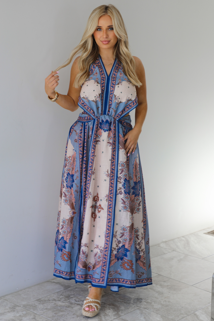 Woman wearing a blue and multi-colored halter tie maxi dress that has a long, flowy skirt with a split hem detail. The photo is taken indoors against a white wall with white marble flooring. 