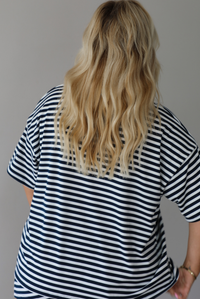 A woman wearing a navy and white horizontally striped short-sleeve oversized tee and skirt with a white drawstring tie at the waist, standing indoors against a white wall. The photo shows the back of the oversized tee. 