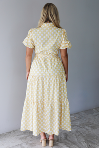 A woman shown from behind wearing a yellow and white polka dot midi dress with puff sleeves and a tie waist and functional button closures along the front, standing indoors against a white wall with white marble flooring. 