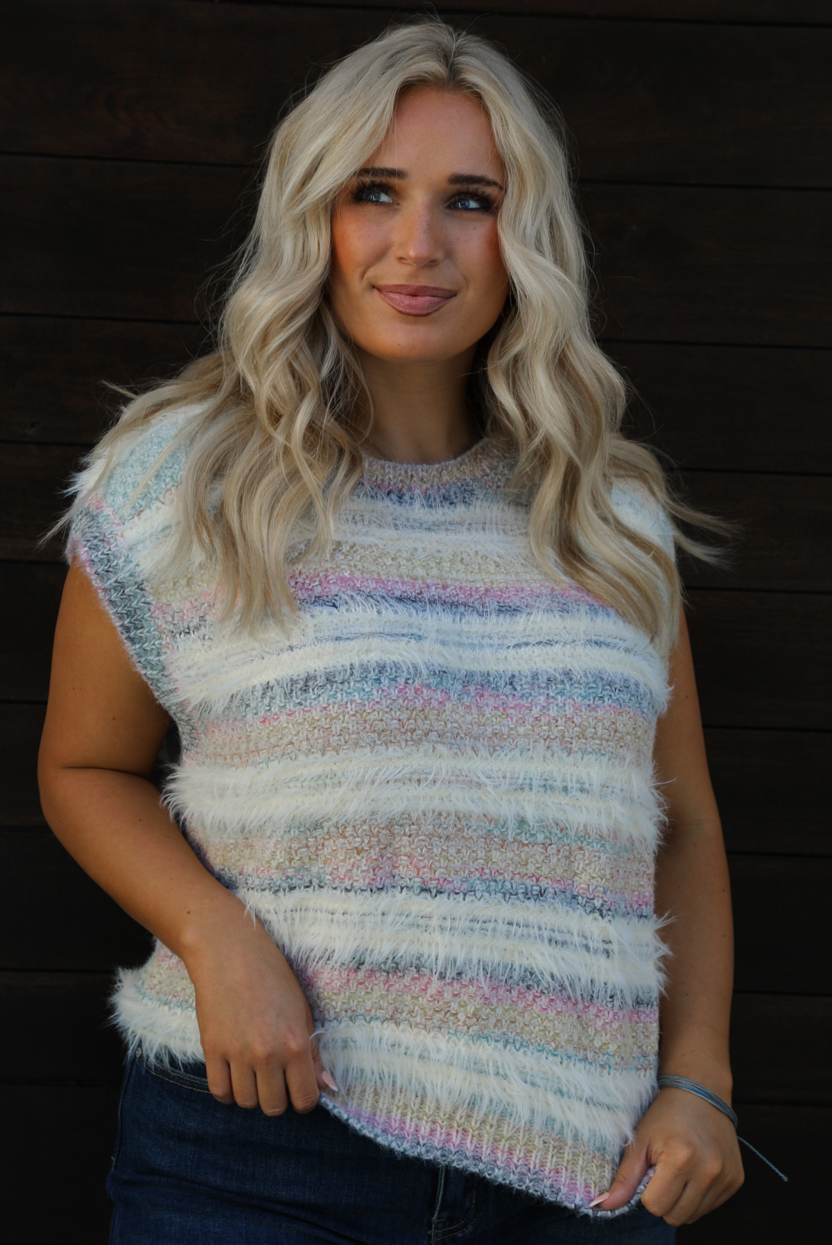 Woman model wearing a soft, striped sweater vest with dark wash denim jeans. The photo showcases a closeup of the sweater vest and the full length. The photo is taken outdoors against a dark brown wooden door. 