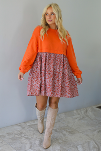 A woman wearing a block style oversized, flowy dress featuring a solid orange bodice and sleeves with a multi-colored floral skirt, paired with tall sand colored boots, standing indoors against a white wall with white marble flooring. 