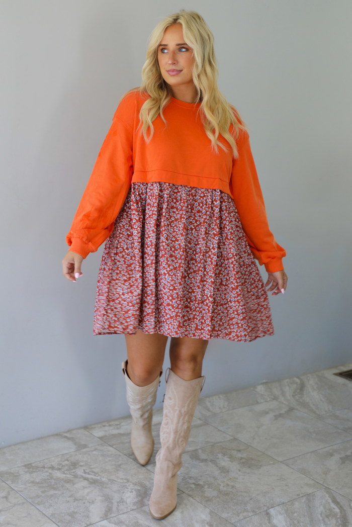 A woman wearing a block style oversized, flowy dress featuring a solid orange bodice and sleeves with a multi-colored floral skirt, paired with tall sand colored boots, standing indoors against a white wall with white marble flooring. 