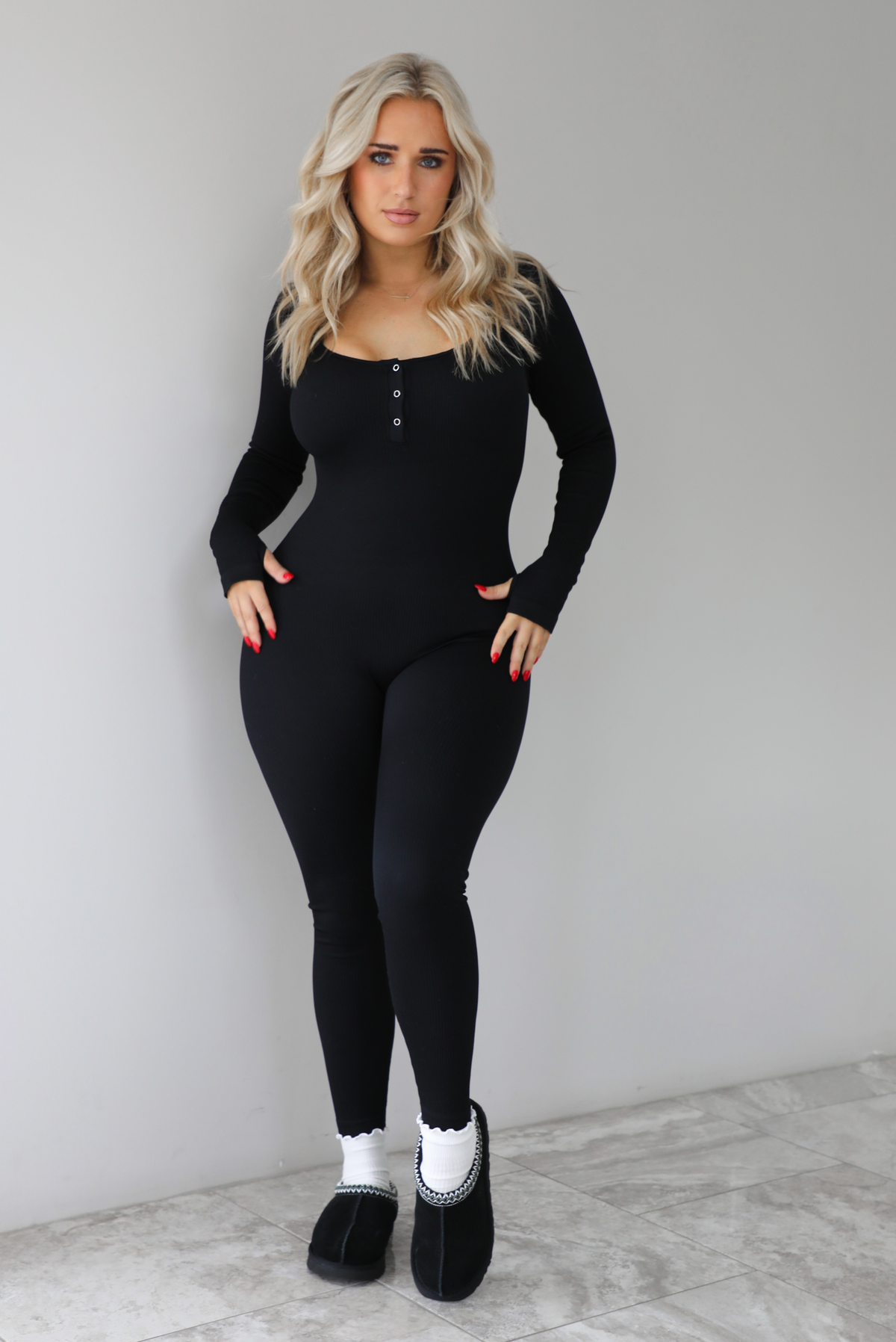 The photo features a model wearing a black fitted jumpsuit with long sleeves that have thumb holes, a rounded neckline with functional button closures, a ribbed texture, and is super stretchy. The jumpsuit is paired with white scrunchie socks and black ugg boots. The photo is taken indoors against a white wall with white marbled flooring. 