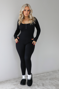 The photo features a model wearing a black fitted jumpsuit with long sleeves that have thumb holes, a rounded neckline with functional button closures, a ribbed texture, and is super stretchy. The jumpsuit is paired with white scrunchie socks and black ugg boots. The photo is taken indoors against a white wall with white marbled flooring. 