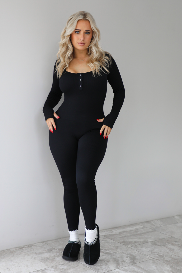 The photo features a model wearing a black fitted jumpsuit with long sleeves that have thumb holes, a rounded neckline with functional button closures, a ribbed texture, and is super stretchy. The jumpsuit is paired with white scrunchie socks and black ugg boots. The photo is taken indoors against a white wall with white marbled flooring. 