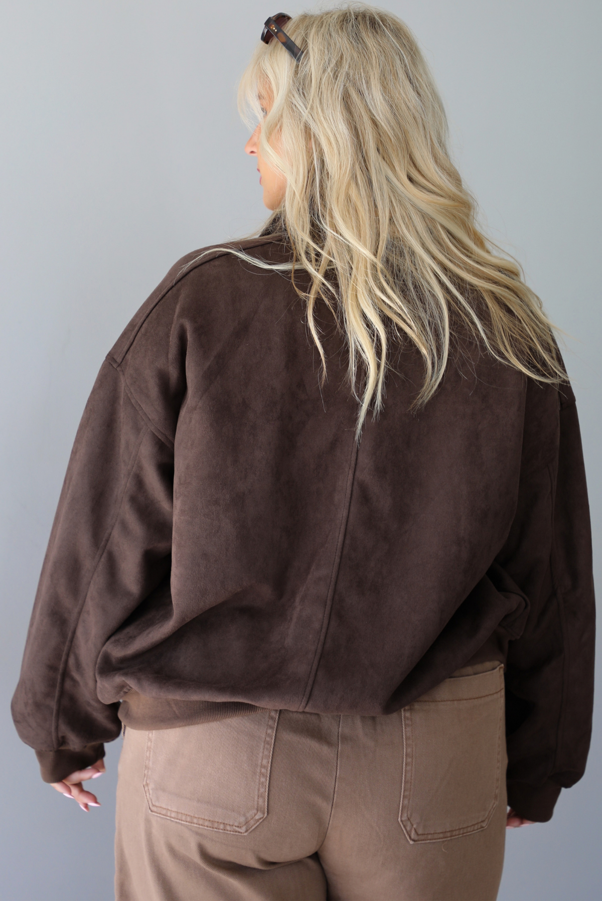 A woman shown from behind wearing a dark brown suede jacket with a white tank and mocha colored mid-to-low rise pants, standing indoors against a white wall.
