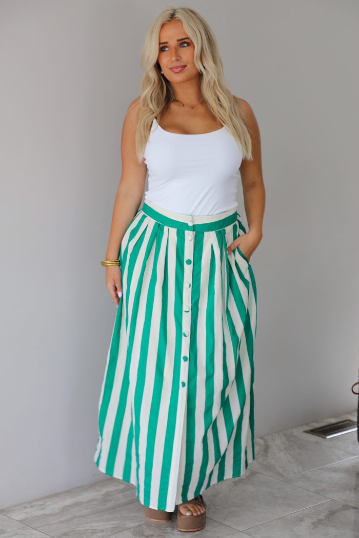 A woman wearing a white tank top and a maxi length cream and green striped skirt,  which has a high waistline, functional pockets on either side, functional button closures in the front, and has a flowy fit, standing indoors against a white wall with white marble flooring. 