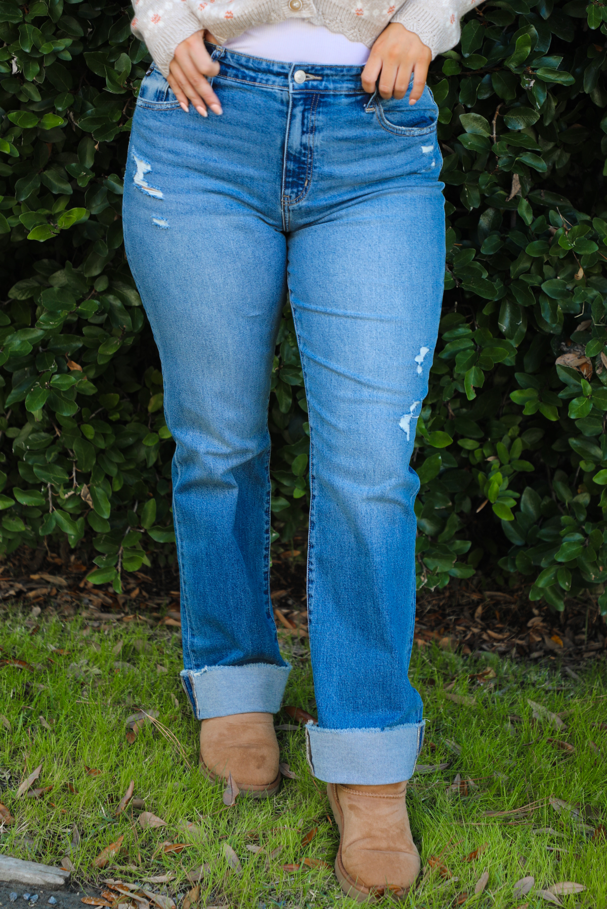 The photo shows a close up of a pair of medium wash denim that have a mid-rise waistline, functional front and back pockets, a button and zipper closure in the front, slightly distressed details on the hips the bottom of the left thigh, and a cuffed hem. The photo is taken outdoors with greenery in the back. 