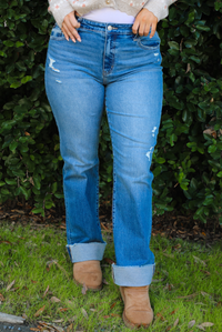 The photo shows a close up of a pair of medium wash denim that have a mid-rise waistline, functional front and back pockets, a button and zipper closure in the front, slightly distressed details on the hips the bottom of the left thigh, and a cuffed hem. The photo is taken outdoors with greenery in the back. 