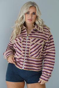 Woman wearing a pink, burgundy, lilac, and sand colored jacket with a charcoal colored two-piece set featuring a cropped sports bra and high waisted short biker shorts. The photo shows the jacket zipped up in the front with the functional pockets in the front. The photo is taken indoors against a white wall.