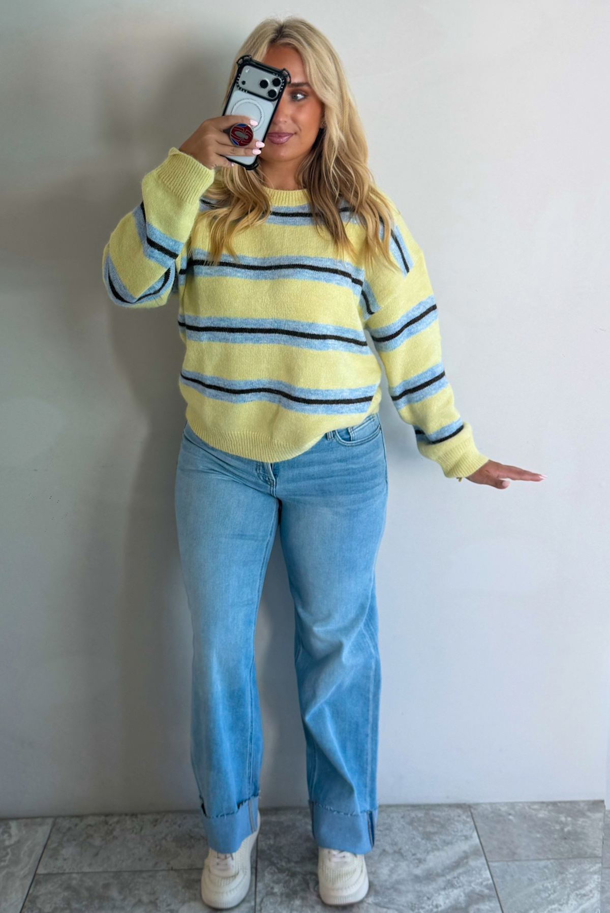 Woman wearing a pastel yellow, blue, and dark brown striped sweater, paired with light wash denim jeans that have a wide leg cut with a cuffed hem, standing indoors against a white wall with white marble flooring, taking a mirror selfie. 