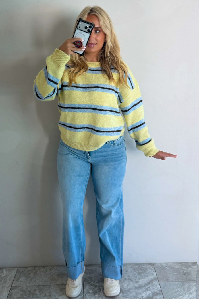 Woman wearing a pastel yellow, blue, and dark brown striped sweater, paired with light wash denim jeans that have a wide leg cut with a cuffed hem, standing indoors against a white wall with white marble flooring, taking a mirror selfie. 