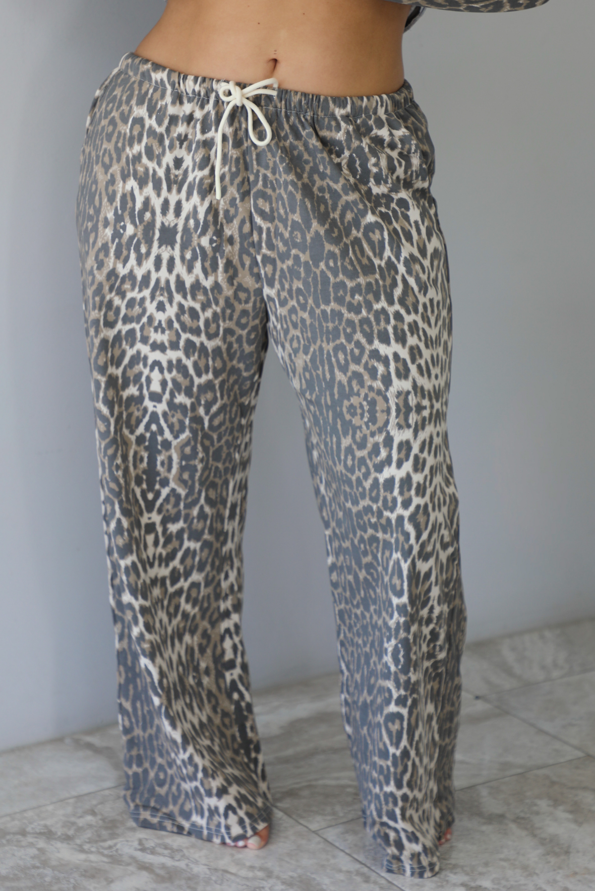 A woman wearing a leopard patterned two piece set featuring an oversized top with a front pocket detail and mid-rise oversized pants, standing indoors against a white wall with white marble flooring. The photo shows a closeup of the pants, which have a drawstring waistline and functional pockets on either side. 