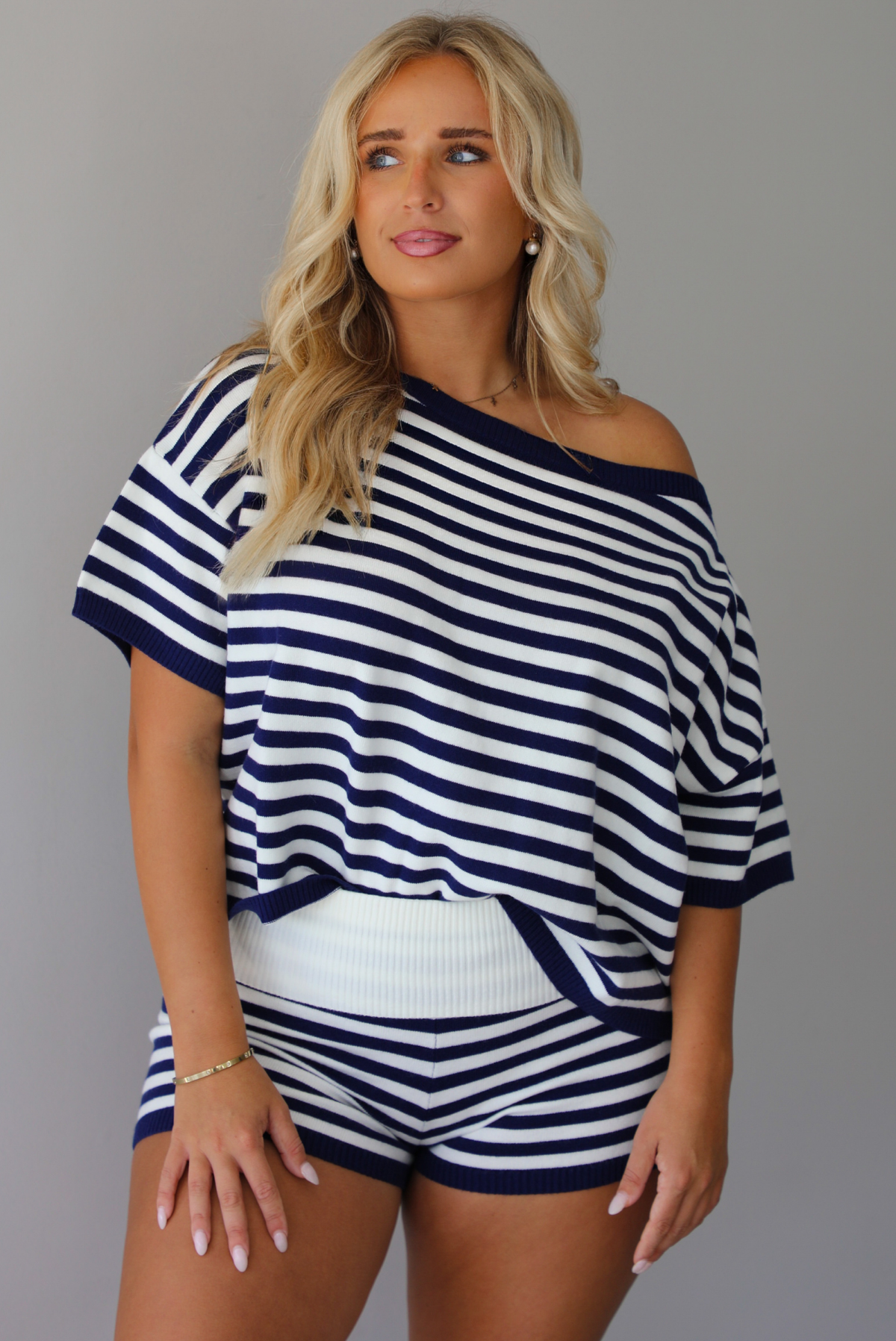 A woman wearing a navy and white horizontally striped off-the-shoulder top and matching striped shorts, standing indoors against a white wall.